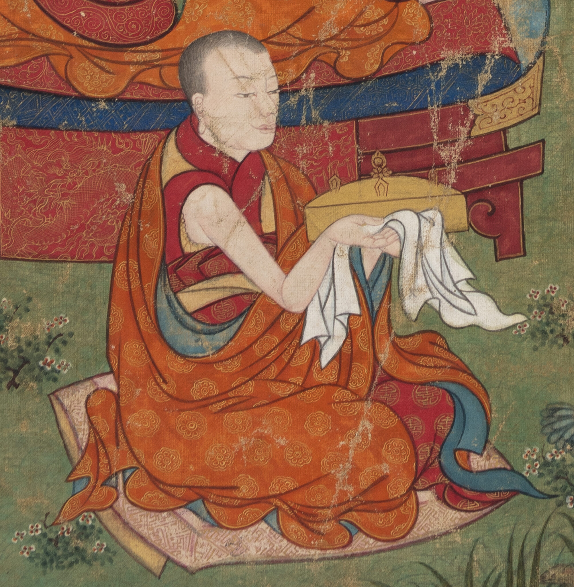 Monk, dressed in saffron brocade robe, kneels and holds forth white prayer scarf and yellow implements