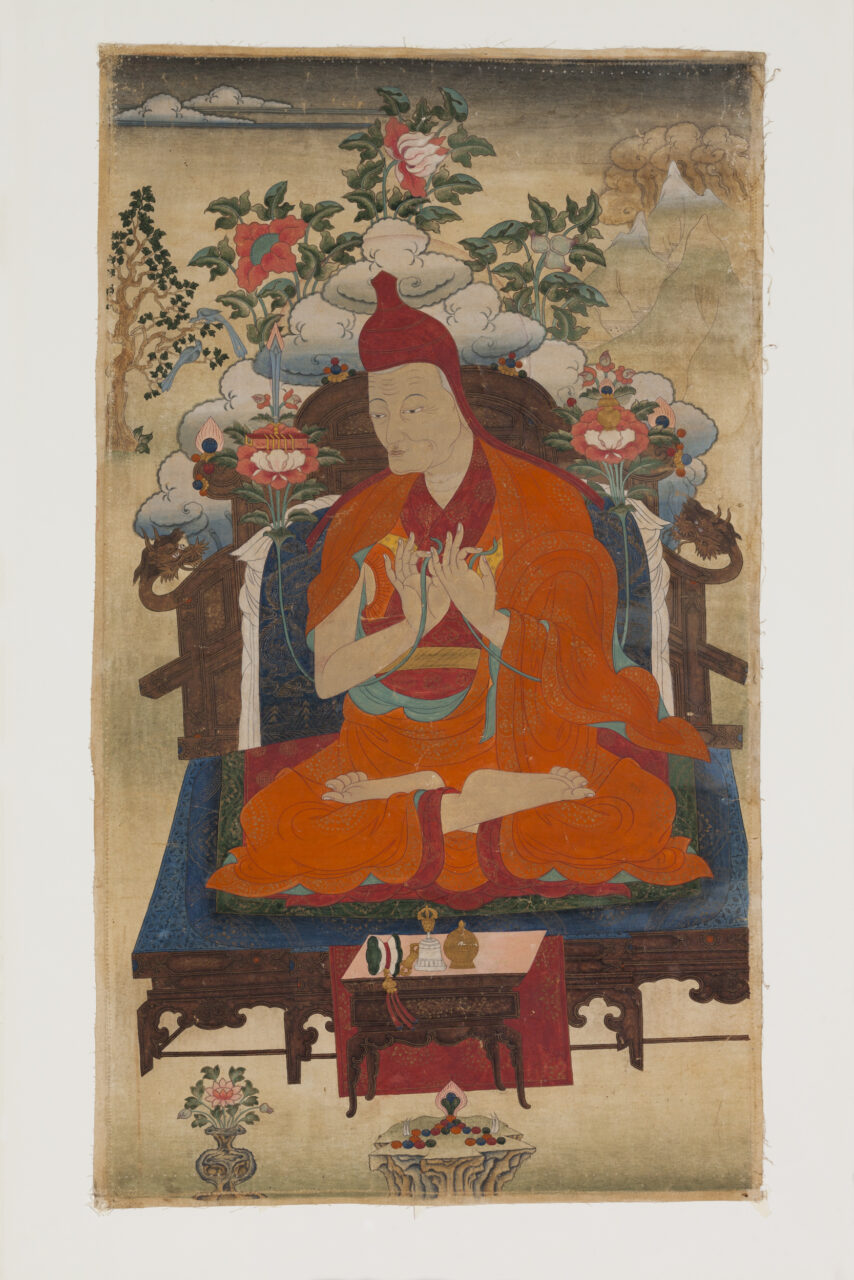 Lama wearing saffron robe seated on blue-cushioned throne with hands posed in mudras at chest