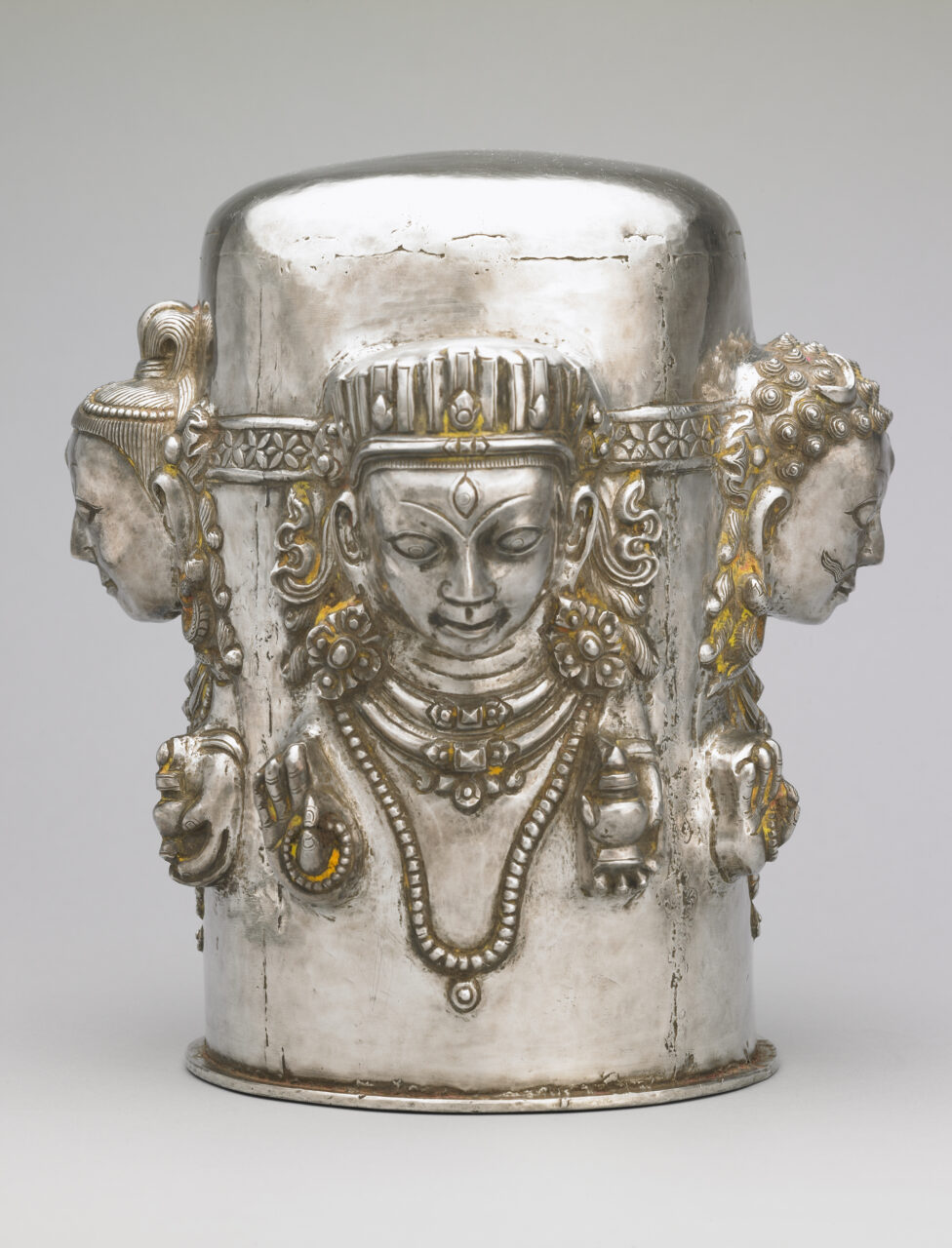 Silver cylindrical object with faces of four figures in relief at cardinal points