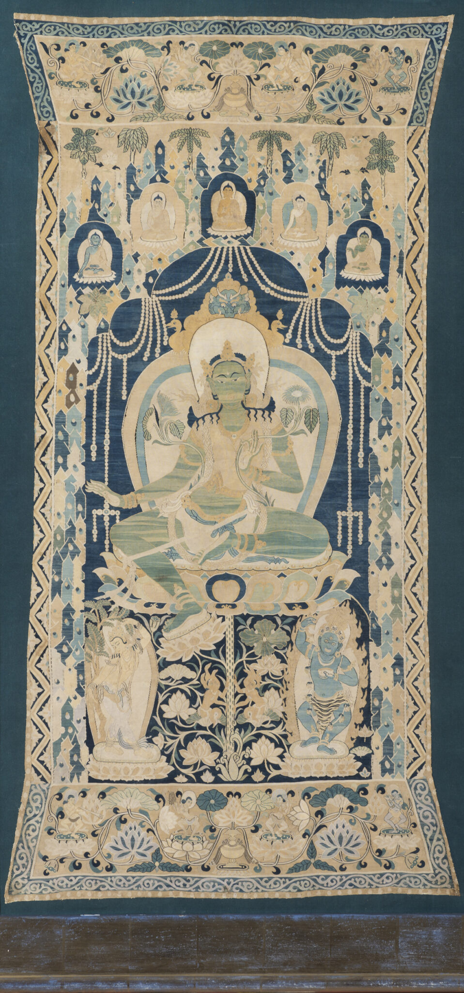 Green-skinned goddess seated underneath arched structure bearing deity portraits and floral motifs in light blues
