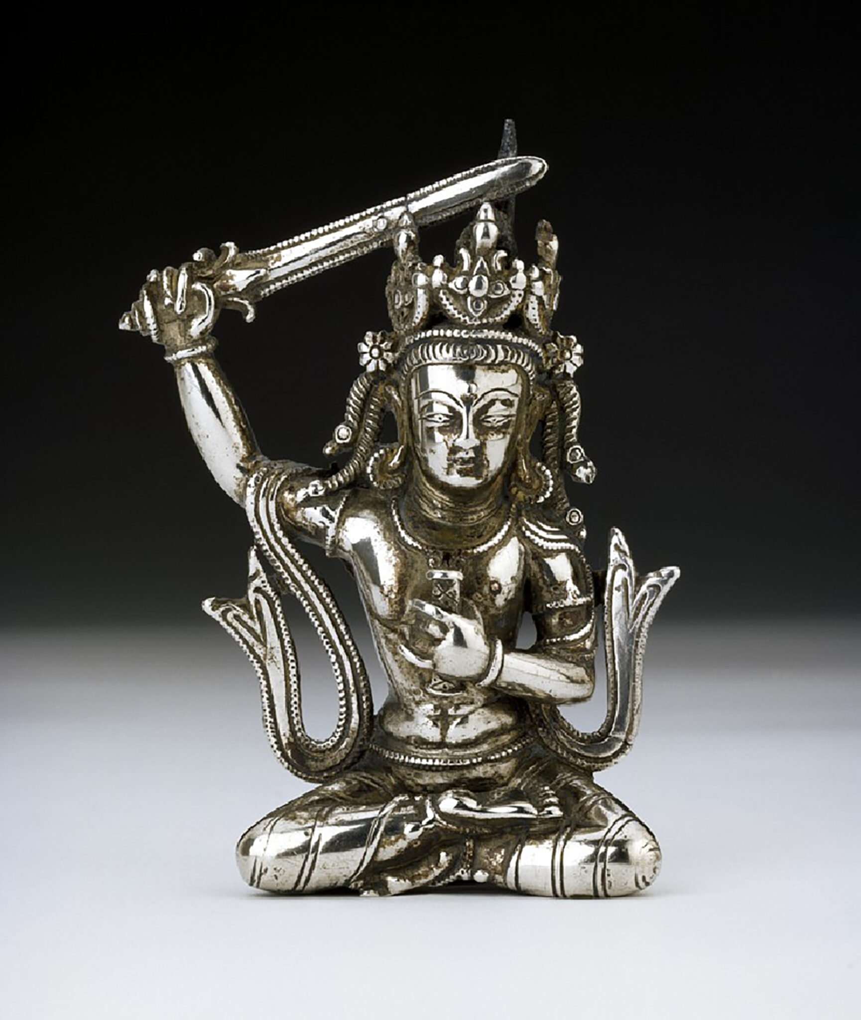 Silver statuette of seated, crowned Bodhisattva, left arm raising sword, with sash billowing about shoulders